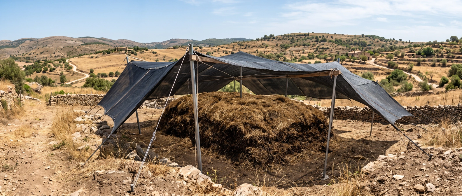 A compost heap capped with a thick straw mulch layer under rigged shade cloth, dry summer hills visible in the background